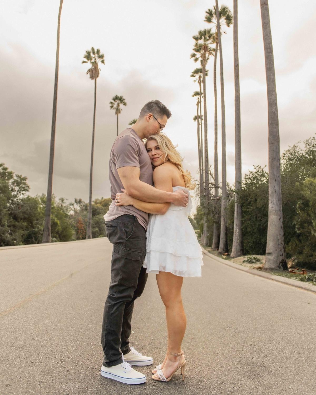 A person kissing a girl on the road with palm trees and a sunset.